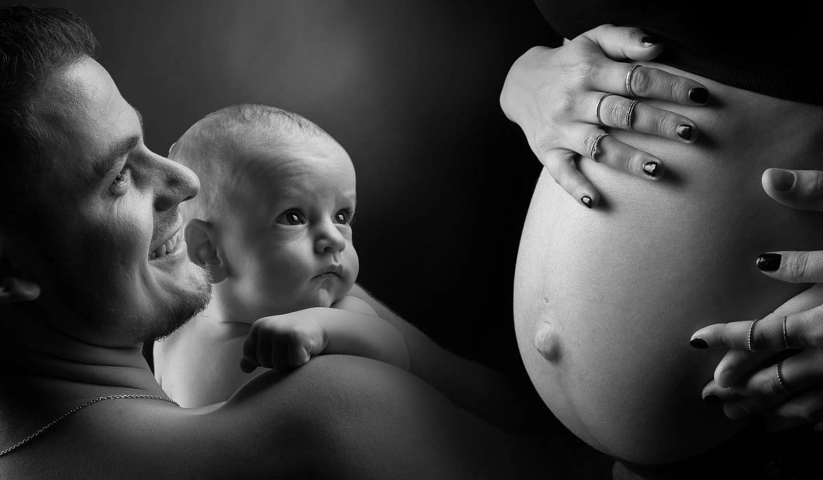 photo noir et blanc prise en studio d'un père qui tient son bébé dans les bras près du ventre d'une femme enceinte
