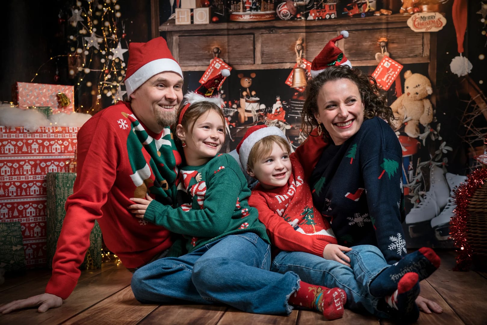 2 parents et 2 enfants sont assis dans un décor de Noël pendant une séance photo. Les 4 portent des pulls de Noël