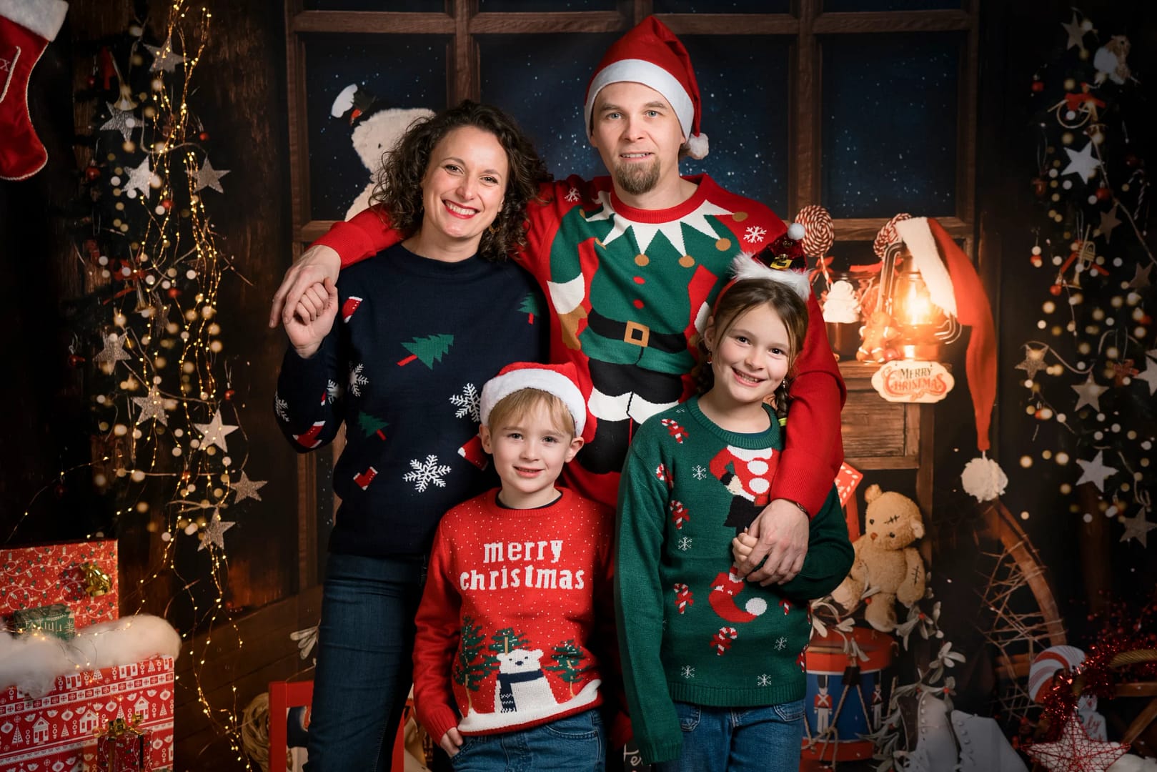 Une famille de 4 personnes portent des pulls de Noël et prennent la pose dans un décor de Noël pendant une séance photo