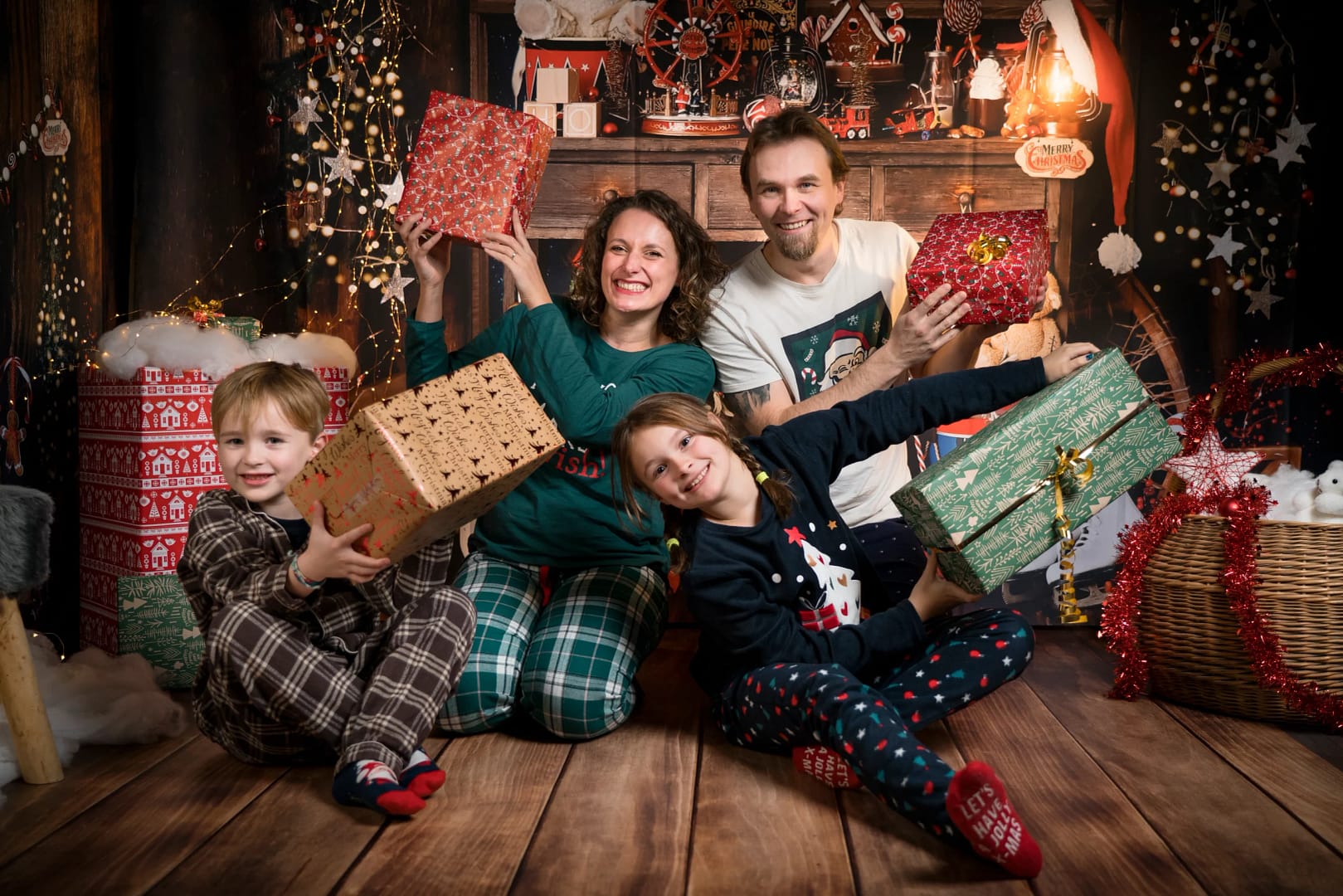 Des parents et leurs 2 enfants font un shooting photo dans un décor de Noël. Ils sont en pyjama et portent des cadeaux dans leurs bras