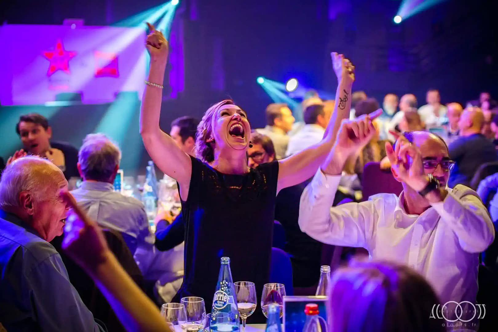 lors d'une soirée d'entreprise dans une grande salle de repas, une femme pousse un cri de joie en se levant de table les bras en l'air. La foule autour d'elle est très animée
