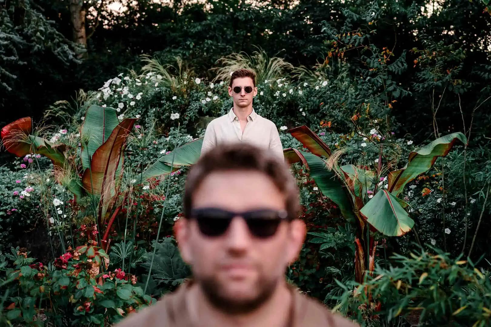 couple d'hommes avec des lunettes de soleil posant dans un parc entourés d'une plantes et de fleurs