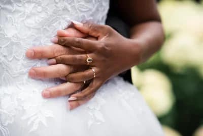 vue rapprochée sur la main d'une mariée portant sa bague de mariage et sa bague de fiançailles. Sa main est posée sur la main de son mari. Les mains sont posées sur le côté de la robe de la mariée