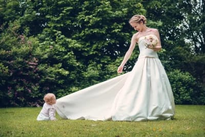 un bébé assis dans l'herbe tire la longue robe d'une mariée qui lui sourit